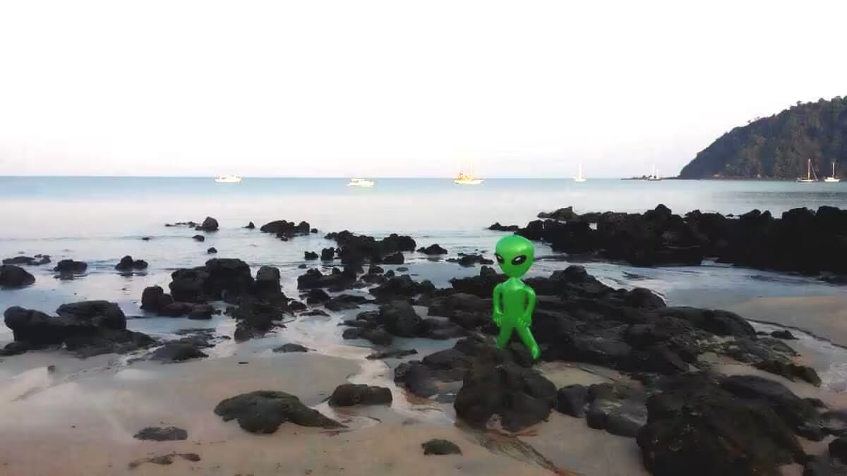 Ein kleiner, grüner Außerirdischer steht zwischen dunklen Felsen am Strand, Hintergrund mit Booten auf dem Meer und einem tohoartigen Landschaftsrand
