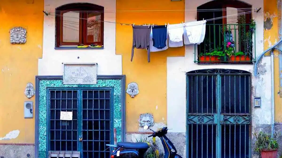 Vor einer gelb-orangen Hausfassade hängen Wäsche und ein Balkon mit Pflanzen, während zwei vergitterte Türen und ein Roller das Stadtbild prägen.